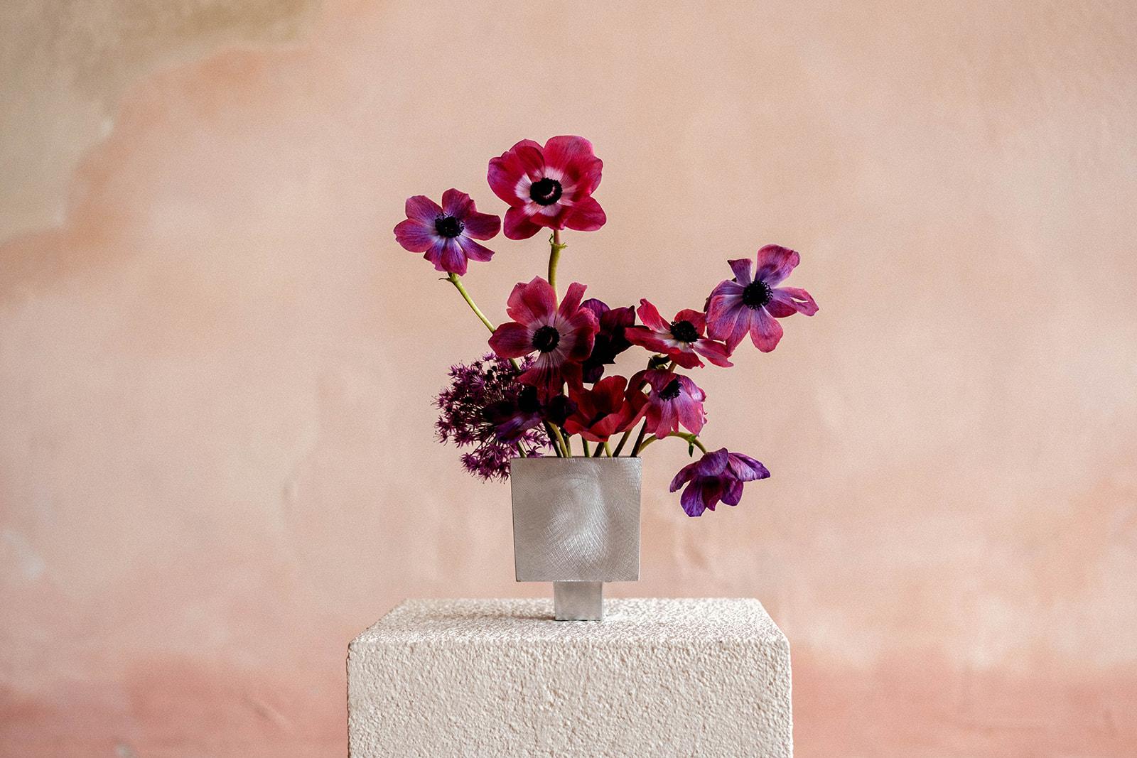 Inky burgundy and violet anemones in a brushed-steel cube against a plaster wall